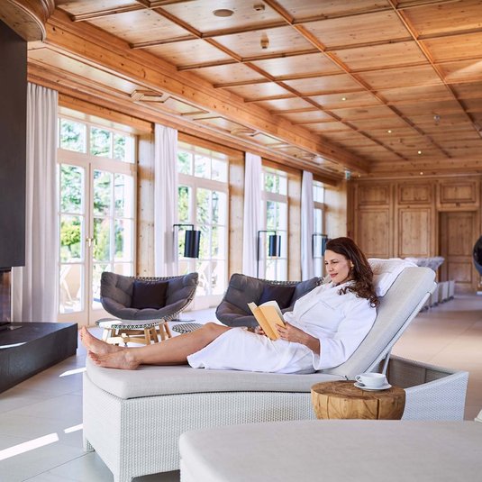 A woman in a white bathrobe lies on a comfortable couch in the relaxation room and reads a book 
